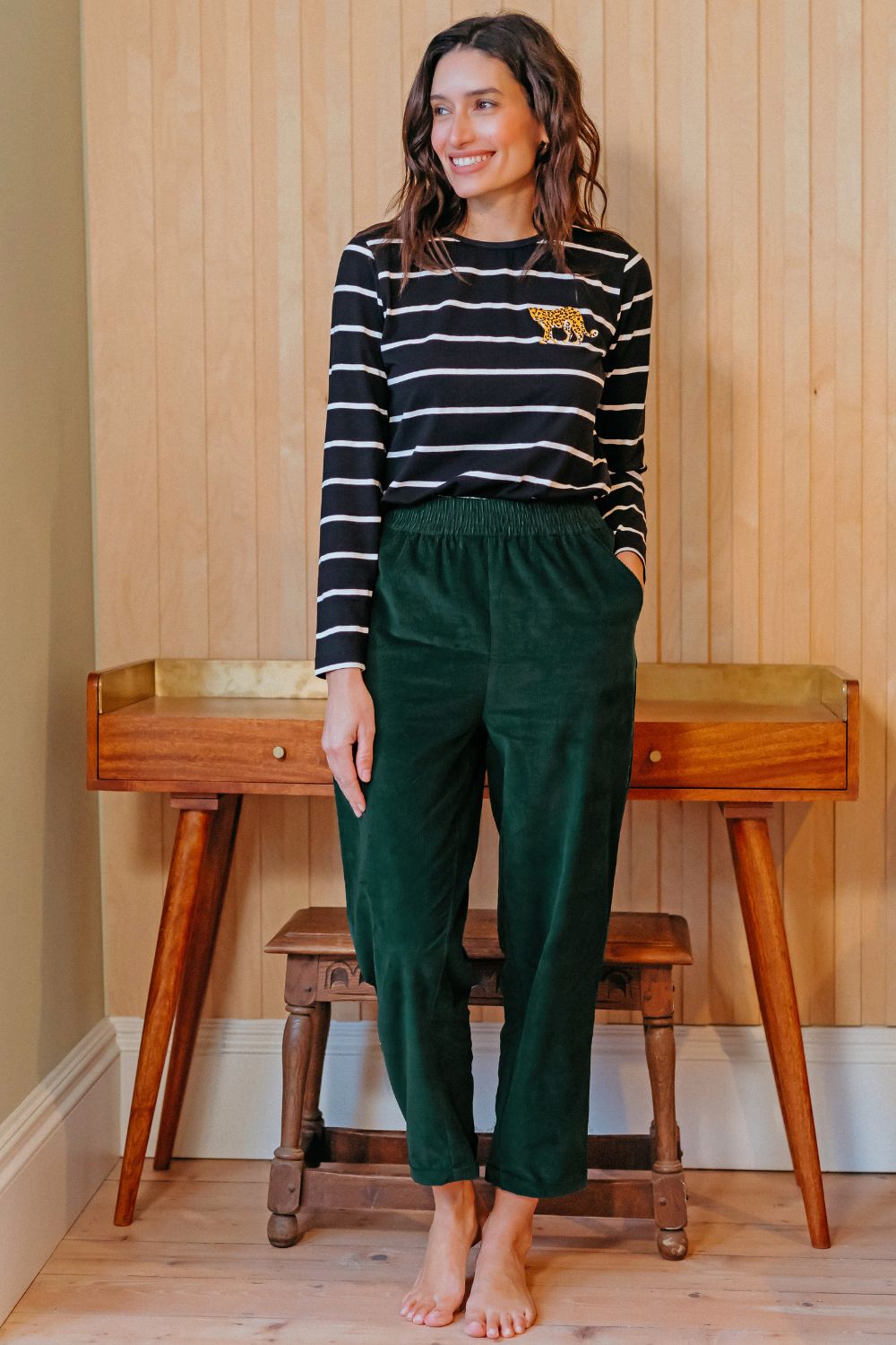 Woman wearing a striped shirt and green pants standing in a room with wooden furniture.