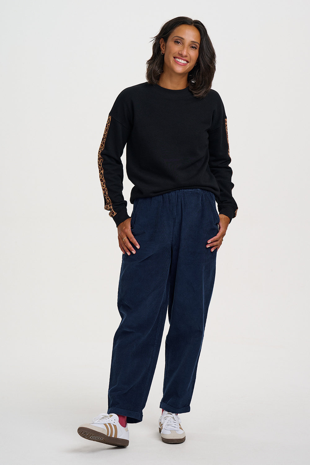 A woman modeling a black crewneck sweatshirt with patterned sleeve details, paired with dark blue corduroy pants and white sneakers, against a light gray studio background.