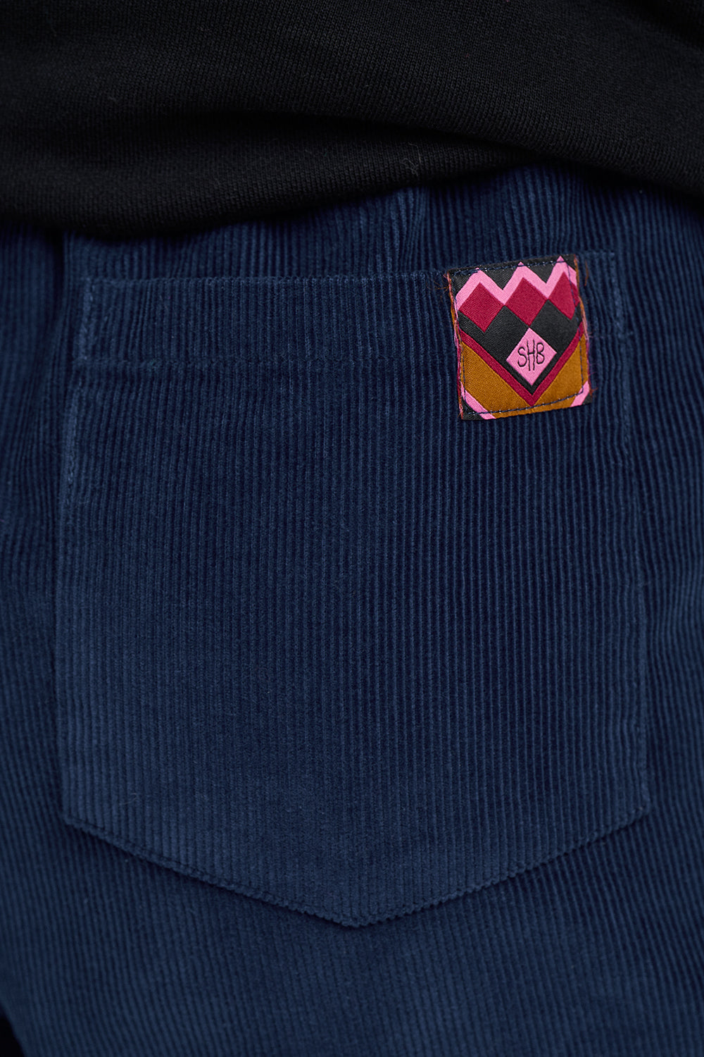 Close-up of a dark blue corduroy pocket with a colorful embroidered patch featuring a mountain-like pattern in pink, yellow, and green, with a small stylized logo in the center.