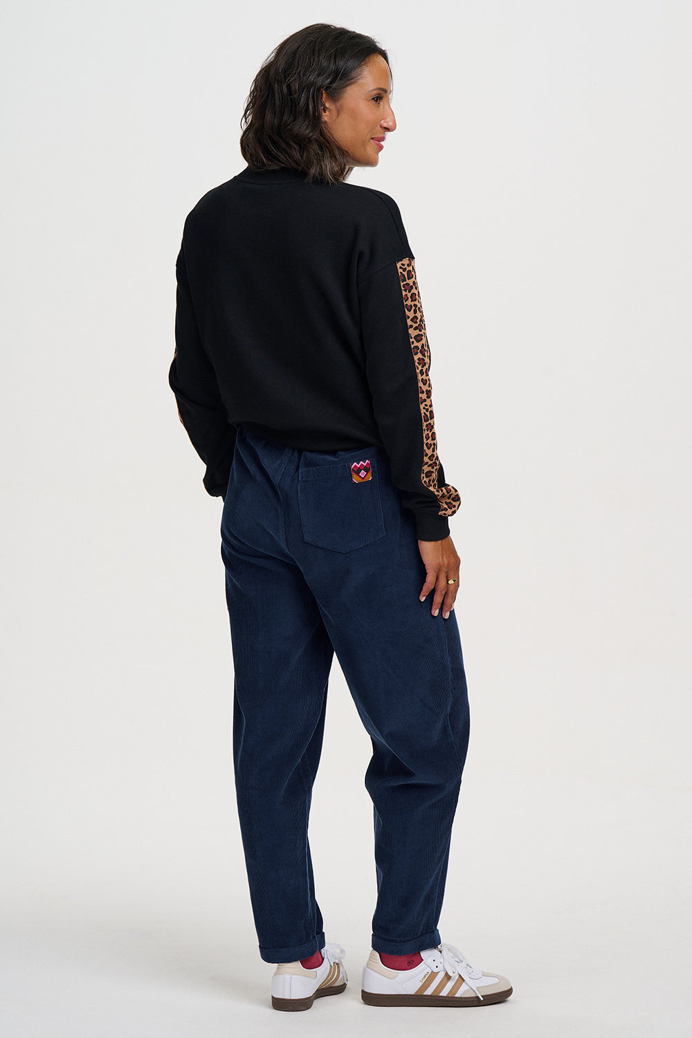 A model shown from the back wearing a black long-sleeve top with a leopard-print sleeve, dark blue pants, and white sneakers with red socks, against a plain white studio background.