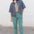 Person wearing a denim shirt over a patterned top with teal pants against a white wall.