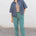 Person wearing a denim shirt over a patterned top with teal pants against a white wall.