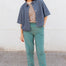Person wearing a denim shirt over a patterned top with teal pants against a white wall.