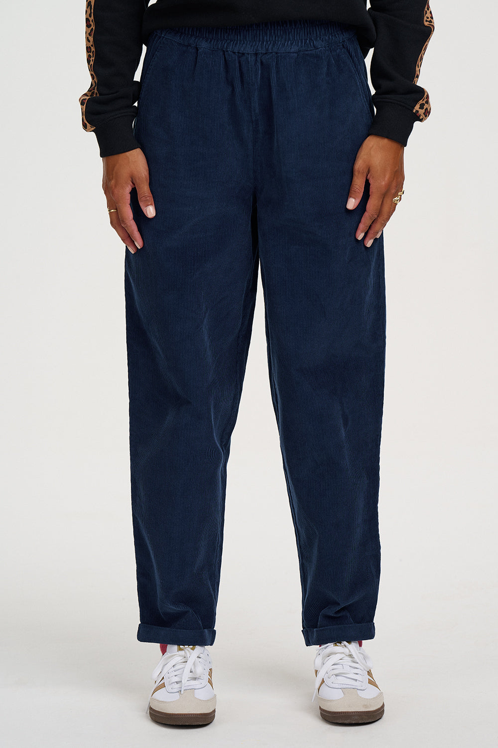 A person wearing dark navy corduroy jogger pants with an elastic waistband and cuffs, paired with white sneakers and a black top with patterned sleeves, shown against a plain light background.