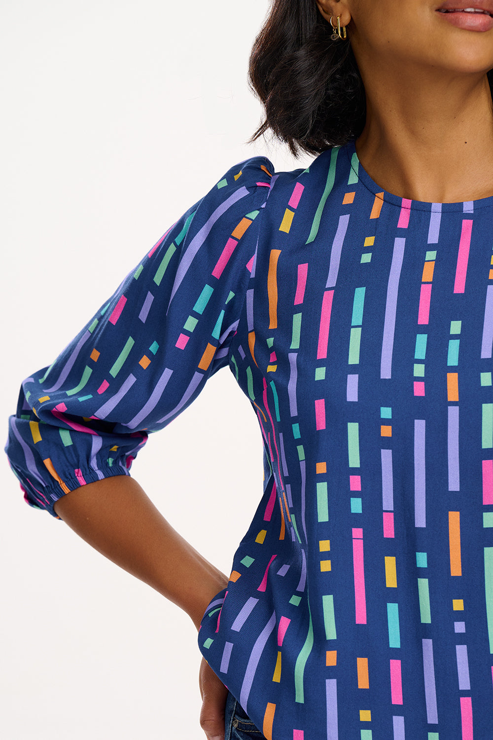 A model wearing a blue blouse with horizontal multicolor lines and 3/4 blouson sleeves.