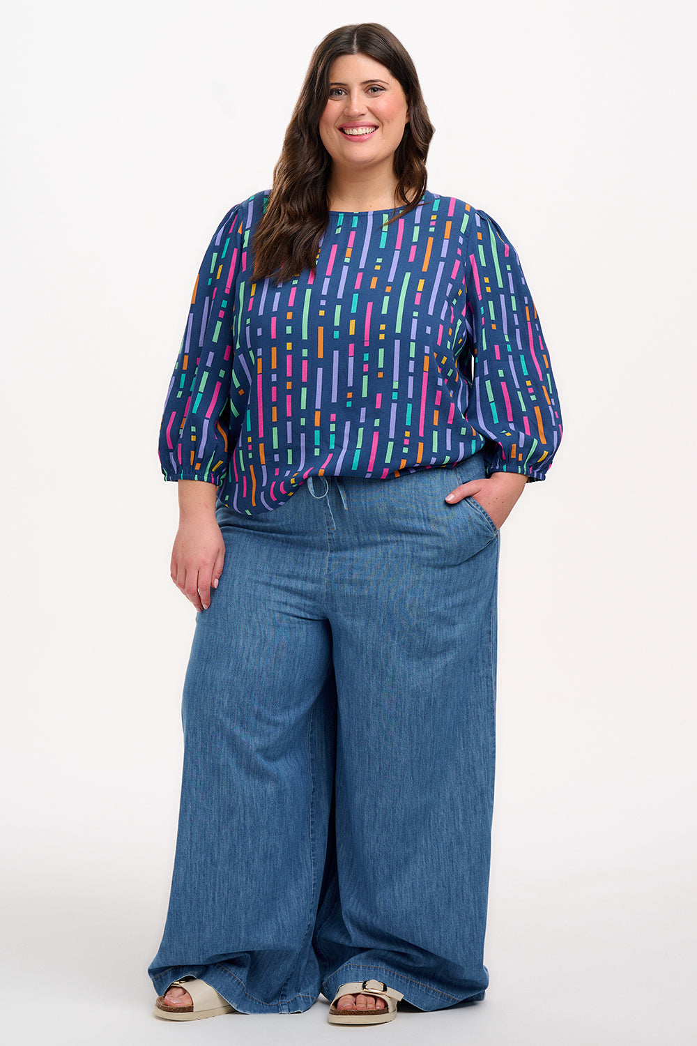 A model wearing a blue blouse with horizontal multicolor lines and 3/4 blouson sleeves.
