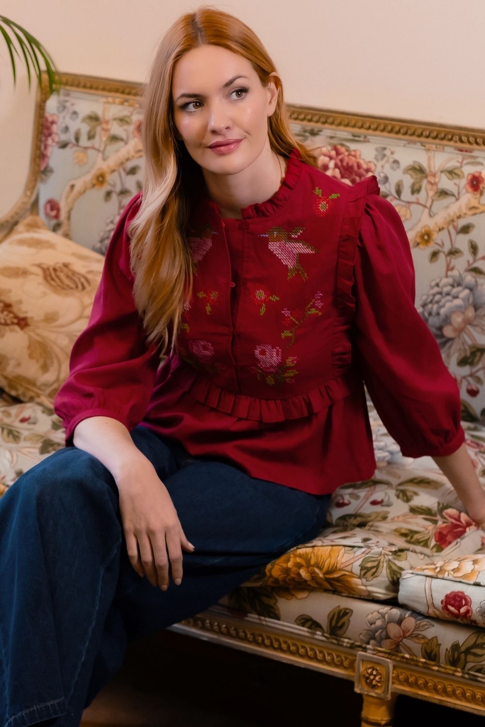 A woman with long red hair sits on an ornate floral sofa wearing a deep red long-sleeve blouse with embroidered birds and flowers, paired with dark blue jeans.