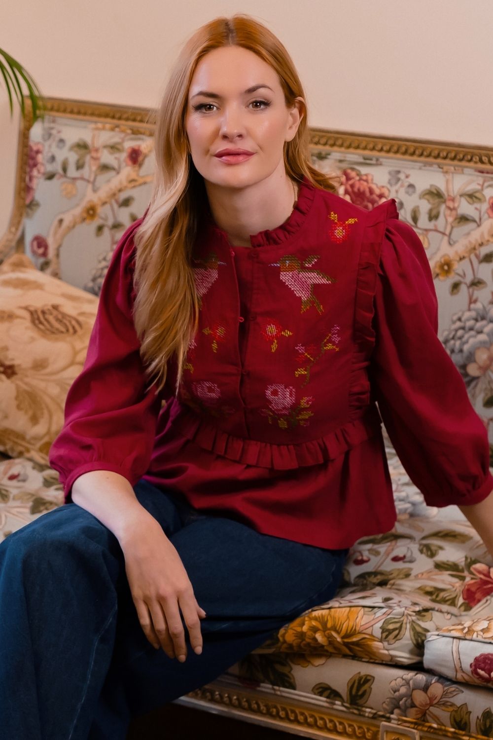 A woman with long light brown hair sits on an ornate floral sofa wearing a deep red long-sleeve blouse with pink and yellow floral embroidery on the chest, paired with dark blue jeans.