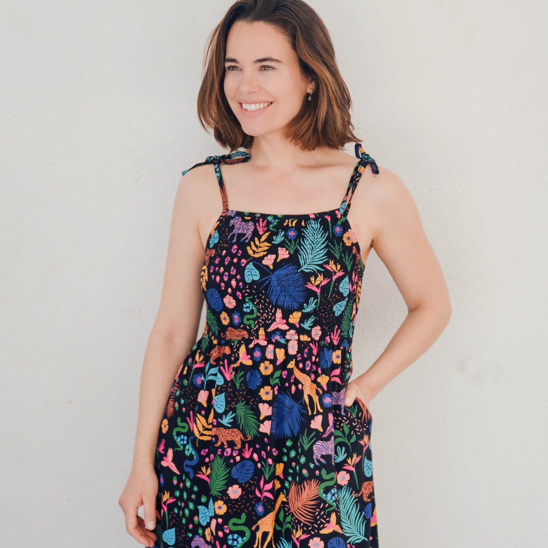 A model wearing a midi length jersey dress with a shirred bodice and tie shoulder straps. The dress has a black base with a colorful jungle spirit design featuring wildlife and foliage.