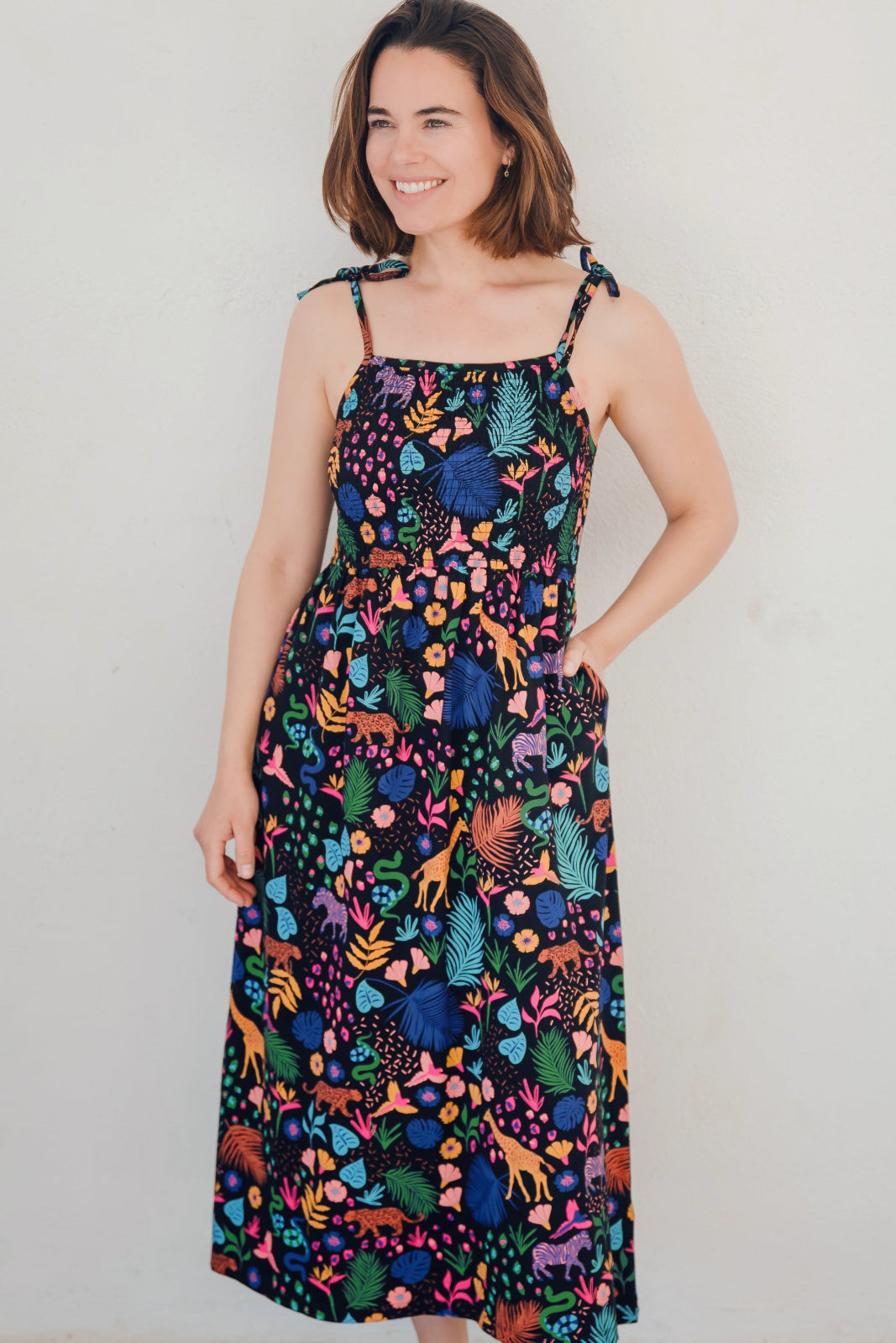 A model wearing a midi length jersey dress with a shirred bodice and tie shoulder straps. The dress has a black base with a colorful jungle spirit design featuring wildlife and foliage.
