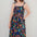 A model wearing a midi length jersey dress with a shirred bodice and tie shoulder straps. The dress has a black base with a colorful jungle spirit design featuring wildlife and foliage.