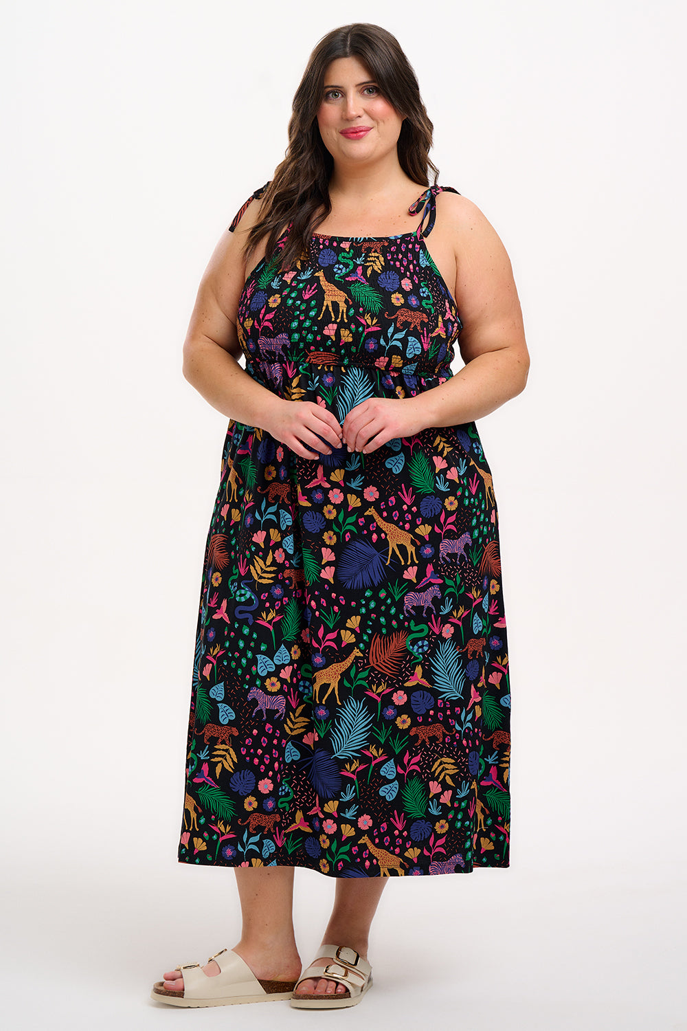 A model wearing a sleeveless, midi-length jersey dress with a multicolored jungle print on a black background, featuring tie shoulder straps.