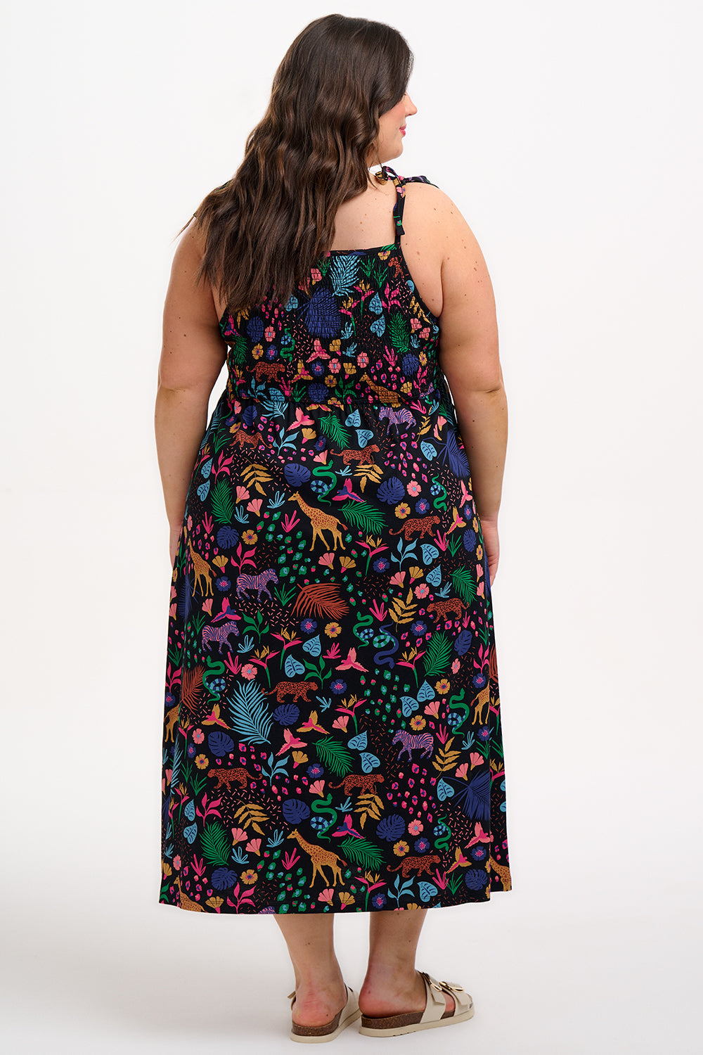A model wearing a sleeveless, midi-length jersey dress with a multicolored jungle print on a black background, featuring tie shoulder straps.