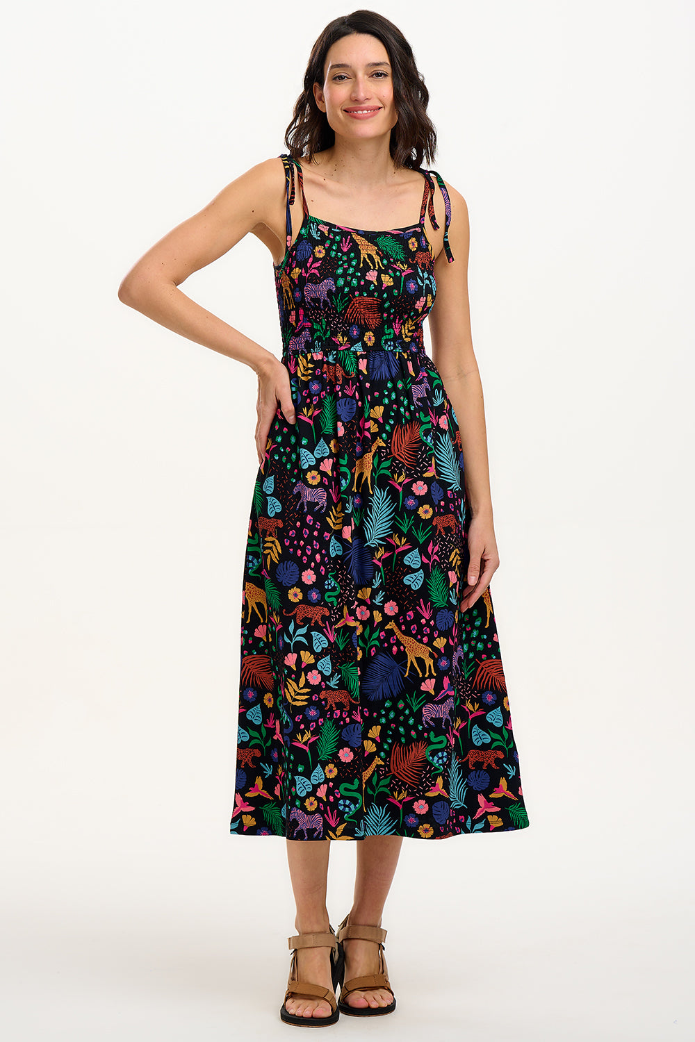 A model wearing a sleeveless, midi-length jersey dress with a multicolored jungle print on a black background, featuring tie shoulder straps.