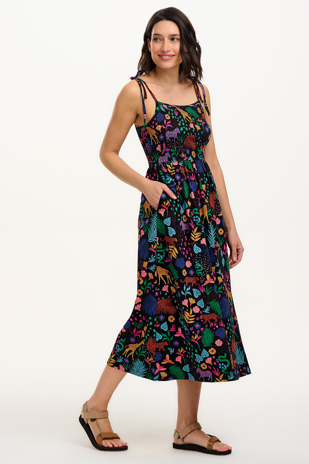A model wearing a sleeveless, midi-length jersey dress with a multicolored jungle print on a black background, featuring tie shoulder straps.