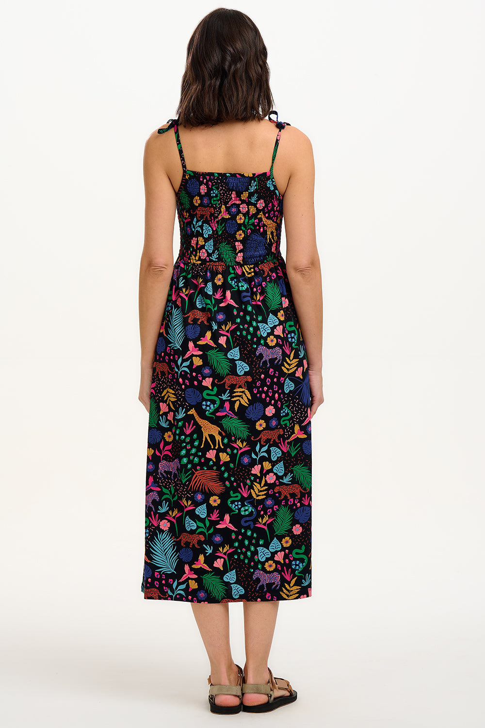 A model wearing a sleeveless, midi-length jersey dress with a multicolored jungle print on a black background, featuring tie shoulder straps.