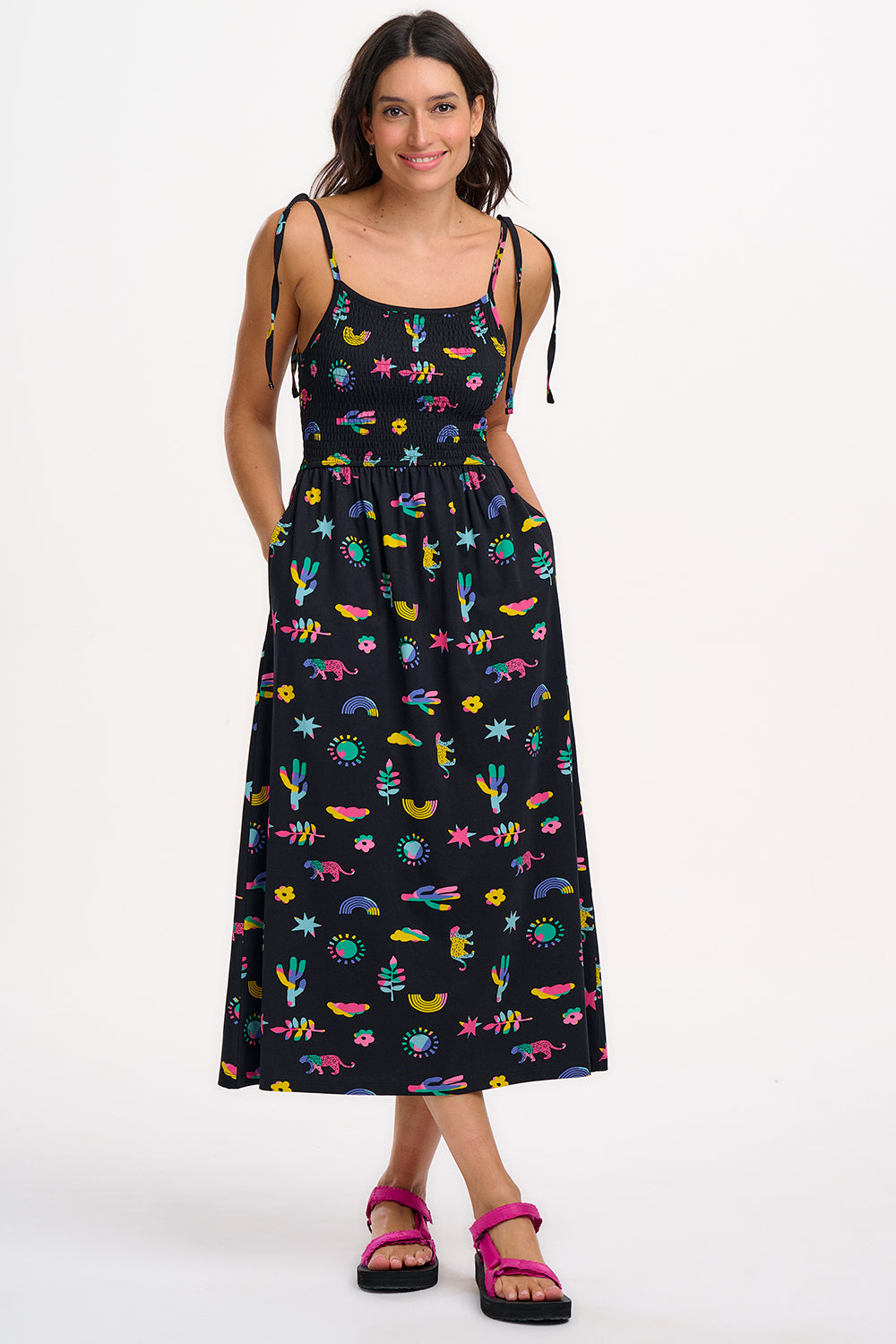 A woman wearing a midi-length black sundress with a colorful pattern, featuring tie shoulder straps and a shirred bodice, standing against a white background.
