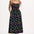 A woman wearing a midi-length black sundress with a colorful pattern, featuring tie shoulder straps and a shirred bodice, standing against a white background.