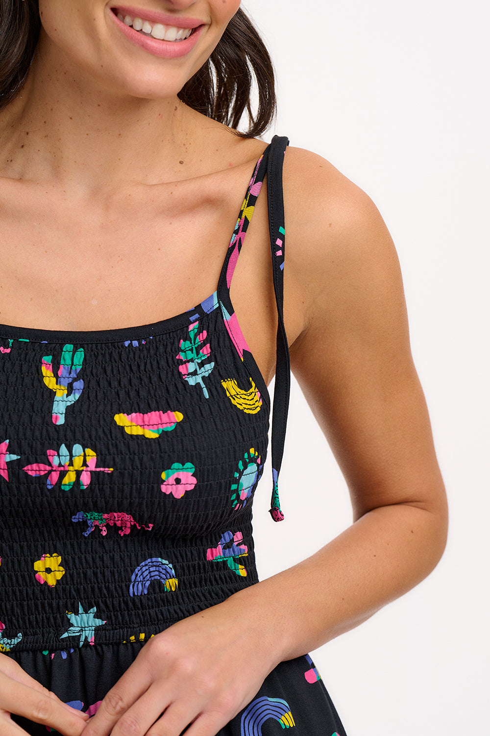 A woman wearing a midi-length black sundress with a colorful pattern, featuring tie shoulder straps and a shirred bodice, standing against a white background.