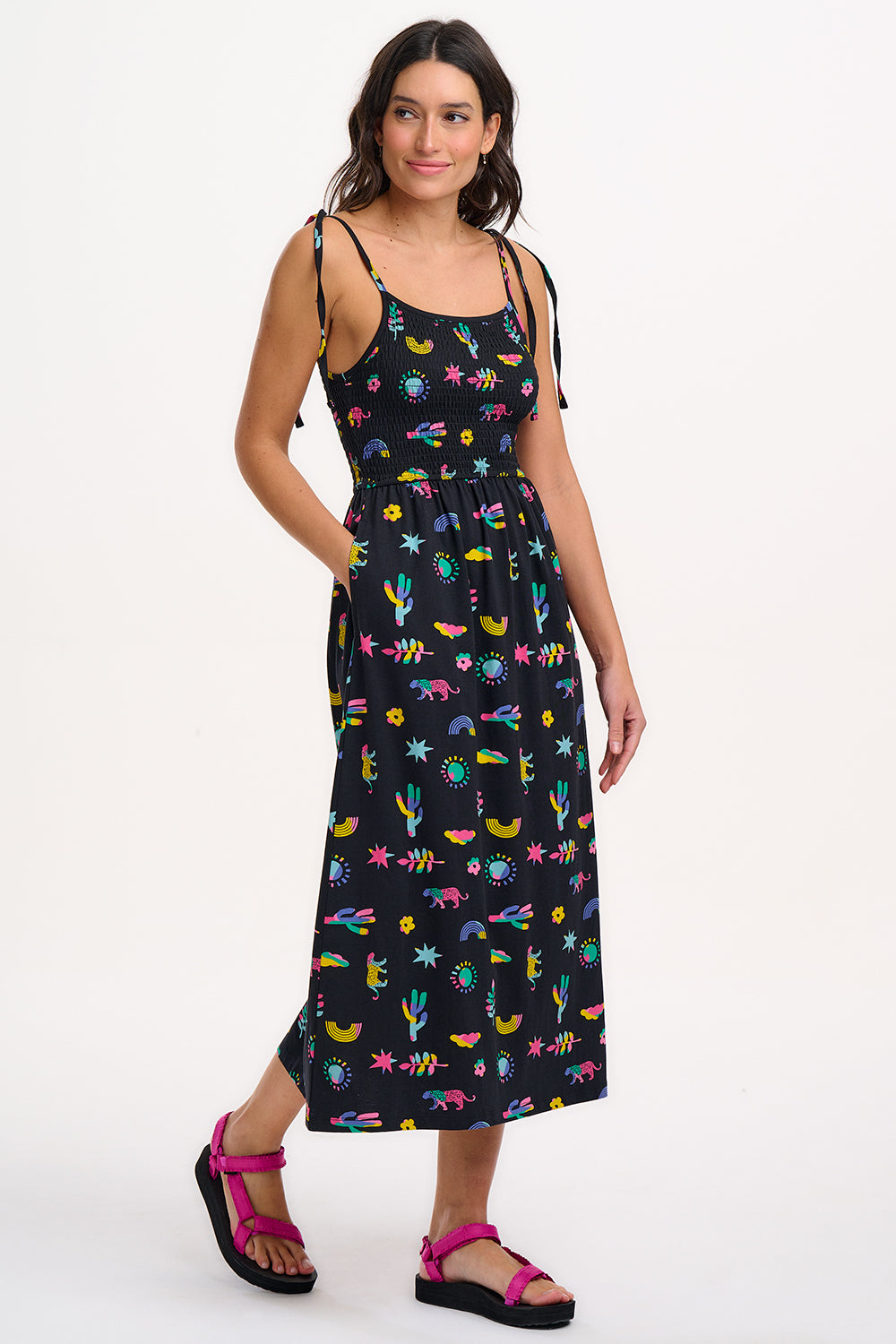 A woman wearing a midi-length black sundress with a colorful pattern, featuring tie shoulder straps and a shirred bodice, standing against a white background.