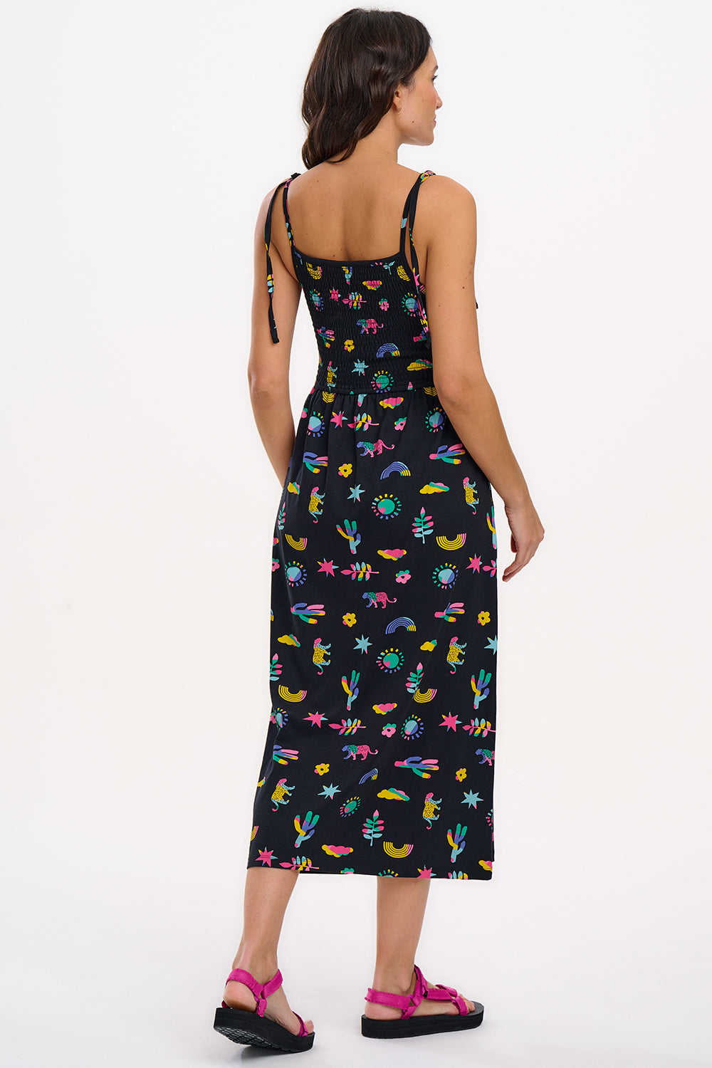 A woman wearing a midi-length black sundress with a colorful pattern, featuring tie shoulder straps and a shirred bodice, standing against a white background.