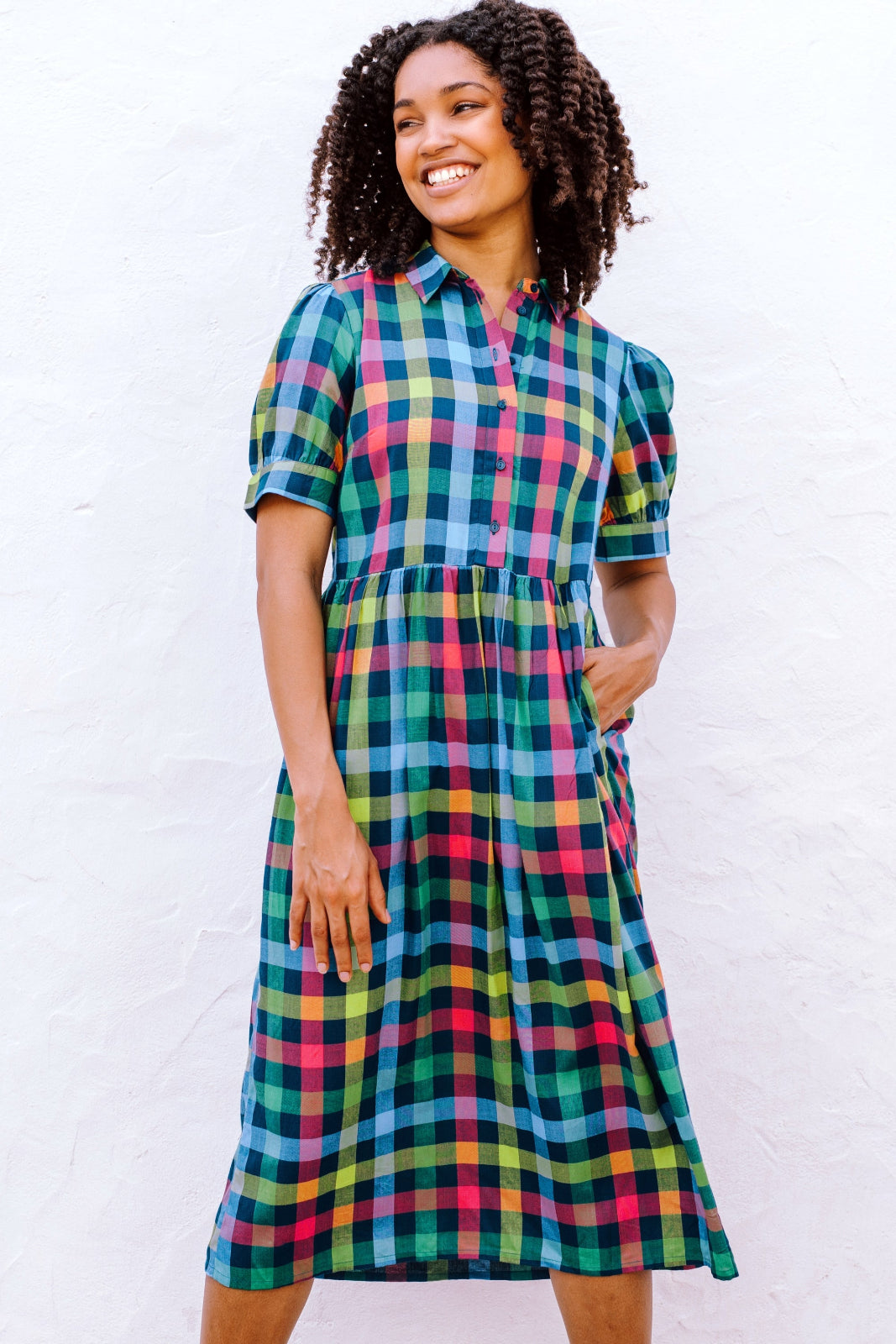 A woman standing and smiling, wearing a midi smock dress with a checkered pattern in shades of pink, green, and blue.