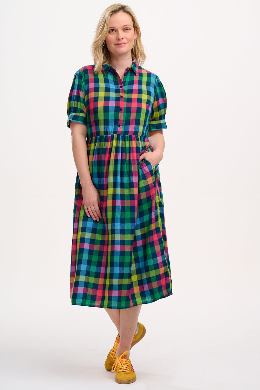 A woman standing and wearing a midi smock dress with a classic shirt bodice and short puff sleeves, featuring a geometric pattern in shades of pink, green, and blue.