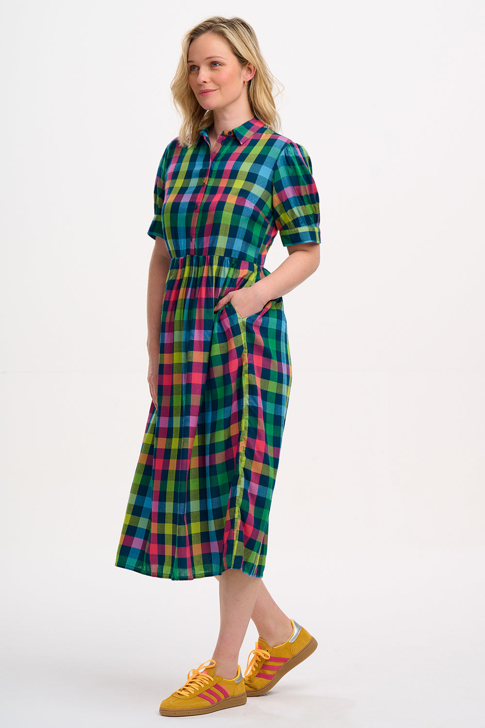 A woman standing and wearing a midi smock dress with a classic shirt bodice and short puff sleeves, featuring a geometric pattern in shades of pink, green, and blue.