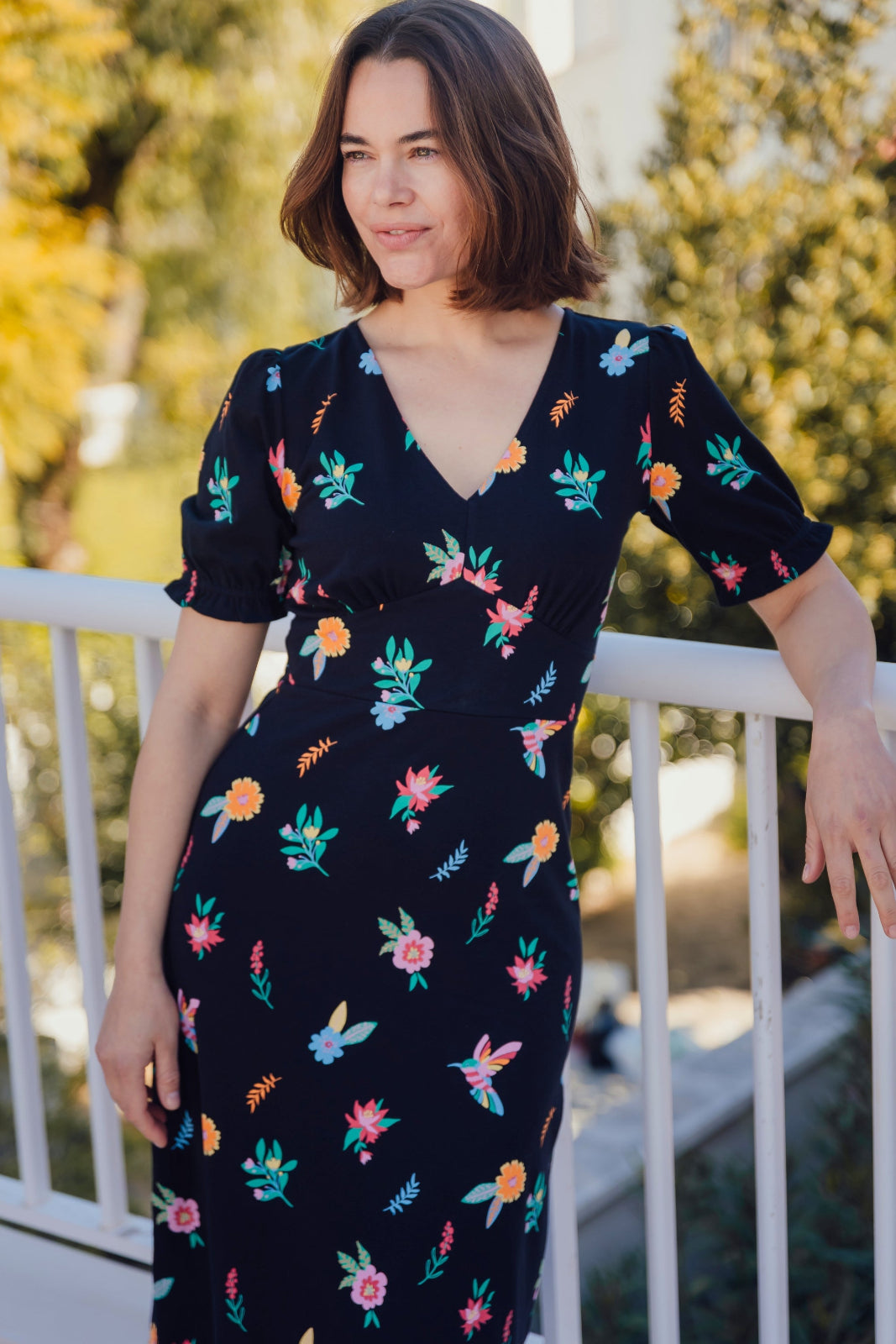 A model wearing a midi length jersey tea dress with a deep V-neck and short frill sleeves, featuring a multicolored hummingbird and flower design on a black base.