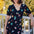 A model wearing a midi length jersey tea dress with a deep V-neck and short frill sleeves, featuring a multicolored hummingbird and flower design on a black base.