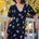 A model wearing a midi length jersey tea dress with a deep V-neck and short frill sleeves, featuring a multicolored hummingbird and flower design on a black base.