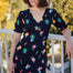 A model wearing a midi length jersey tea dress with a deep V-neck and short frill sleeves, featuring a multicolored hummingbird and flower design on a black base.