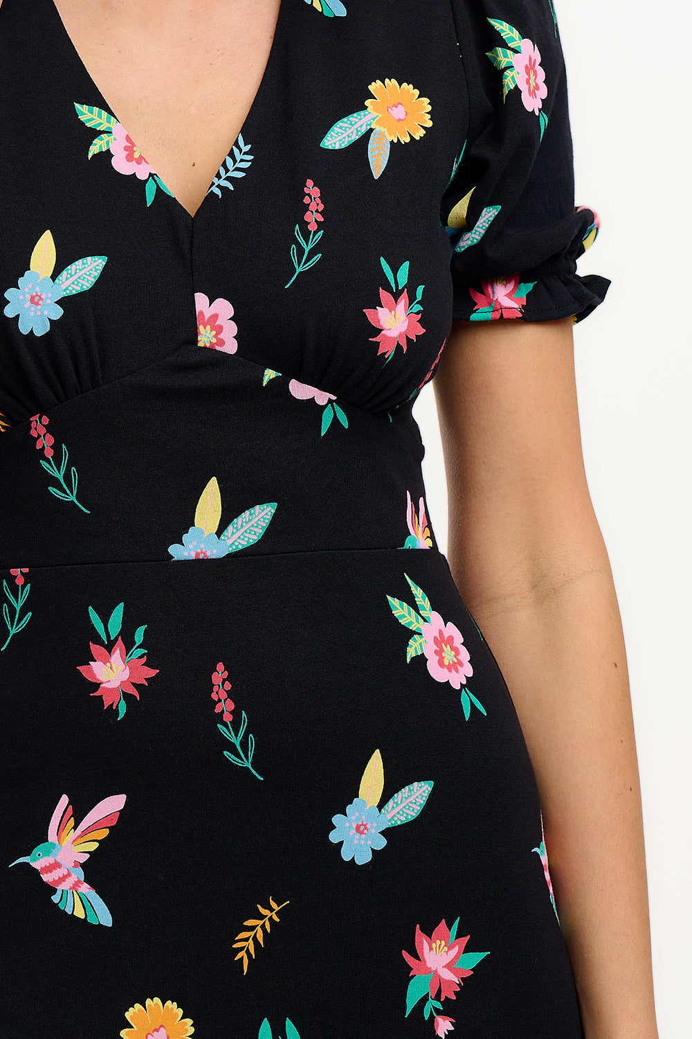 A model wearing a midi length jersey dress with a deep V-neck and short frill sleeves. The dress has a black base with a colorful hummingbird and garden design.