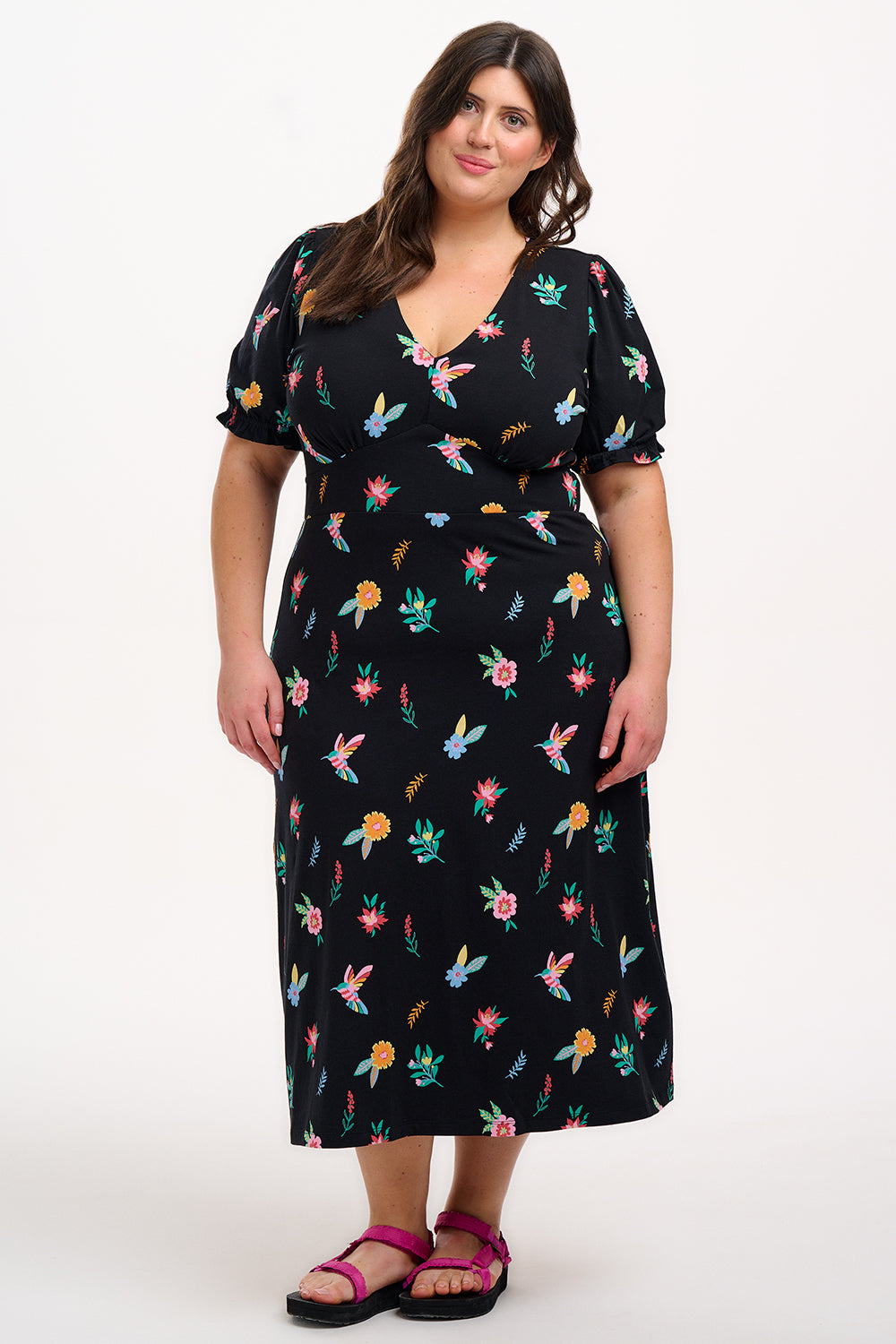 A model wearing a midi length jersey dress with a deep V-neck and short frill sleeves. The dress has a black base with a colorful hummingbird and garden design.