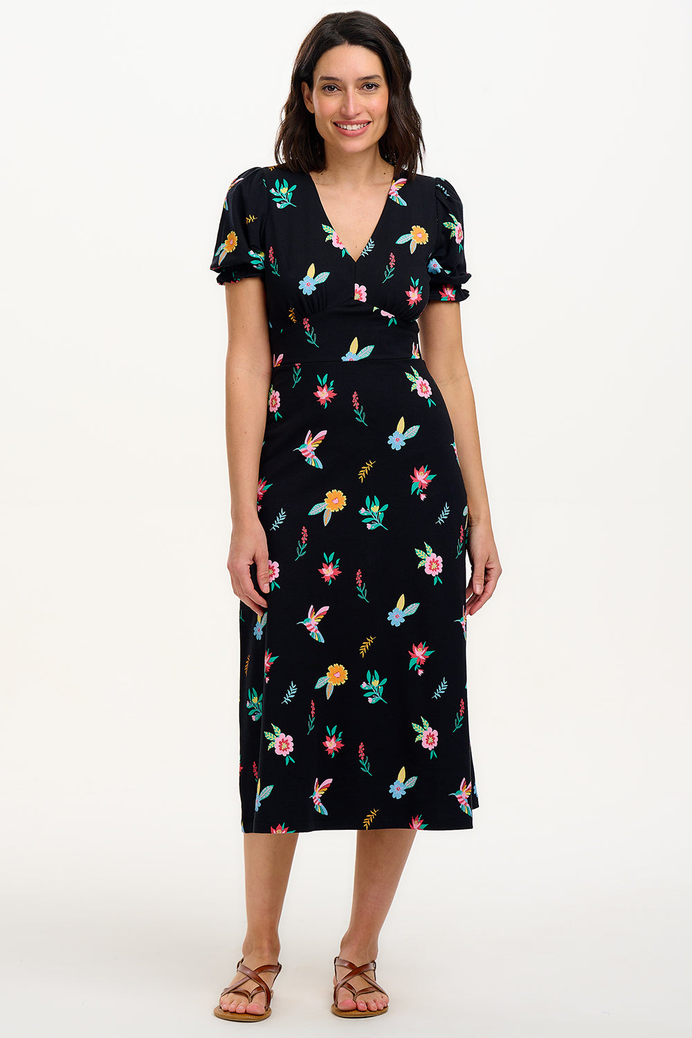 A model wearing a midi length jersey dress with a deep V-neck and short frill sleeves. The dress has a black base with a colorful hummingbird and garden design.