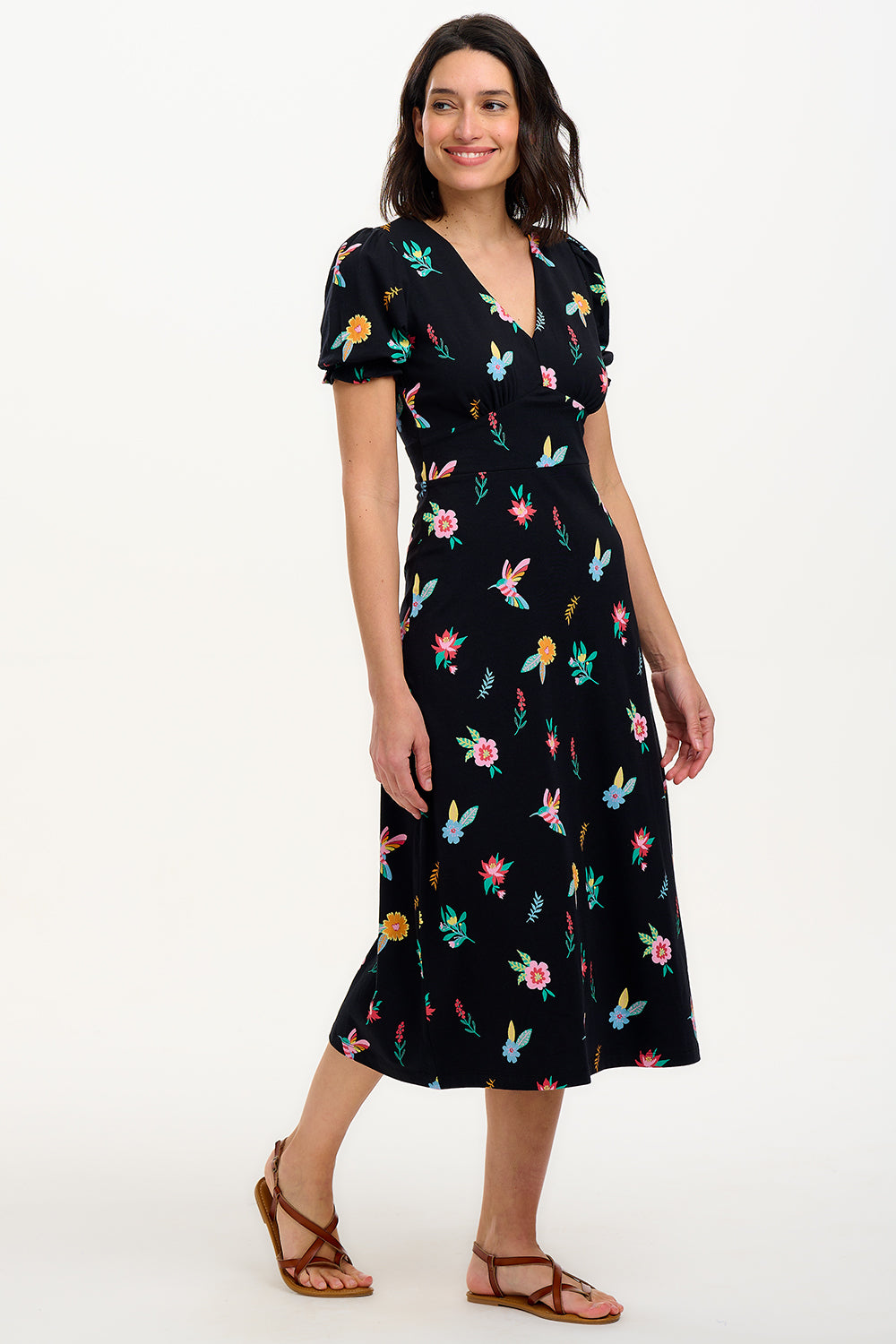 A model wearing a midi length jersey dress with a deep V-neck and short frill sleeves. The dress has a black base with a colorful hummingbird and garden design.
