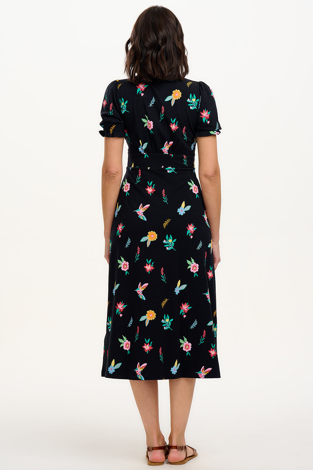 A model wearing a midi length jersey dress with a deep V-neck and short frill sleeves. The dress has a black base with a colorful hummingbird and garden design.