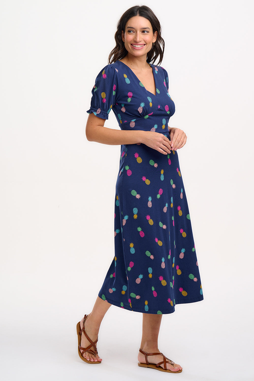 A navy midi tea dress with a multicolor pineapple print, featuring a deep V-neck and short frill sleeves, worn by a model standing against a white background.