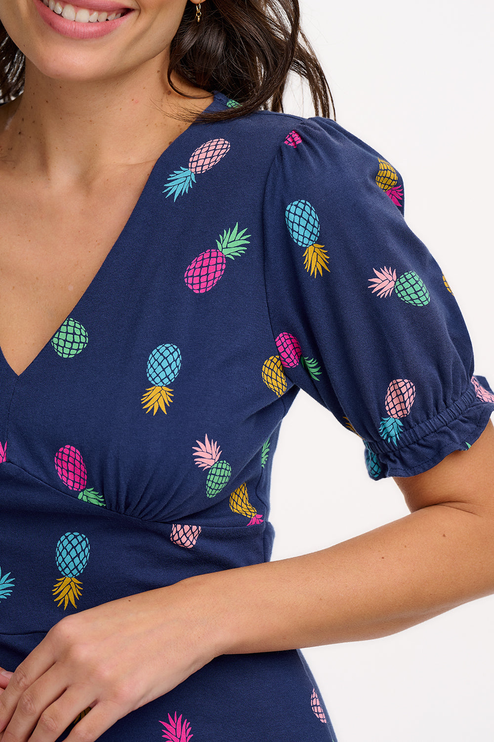 A navy midi tea dress with a multicolor pineapple print, featuring a deep V-neck and short frill sleeves, worn by a model standing against a white background.
