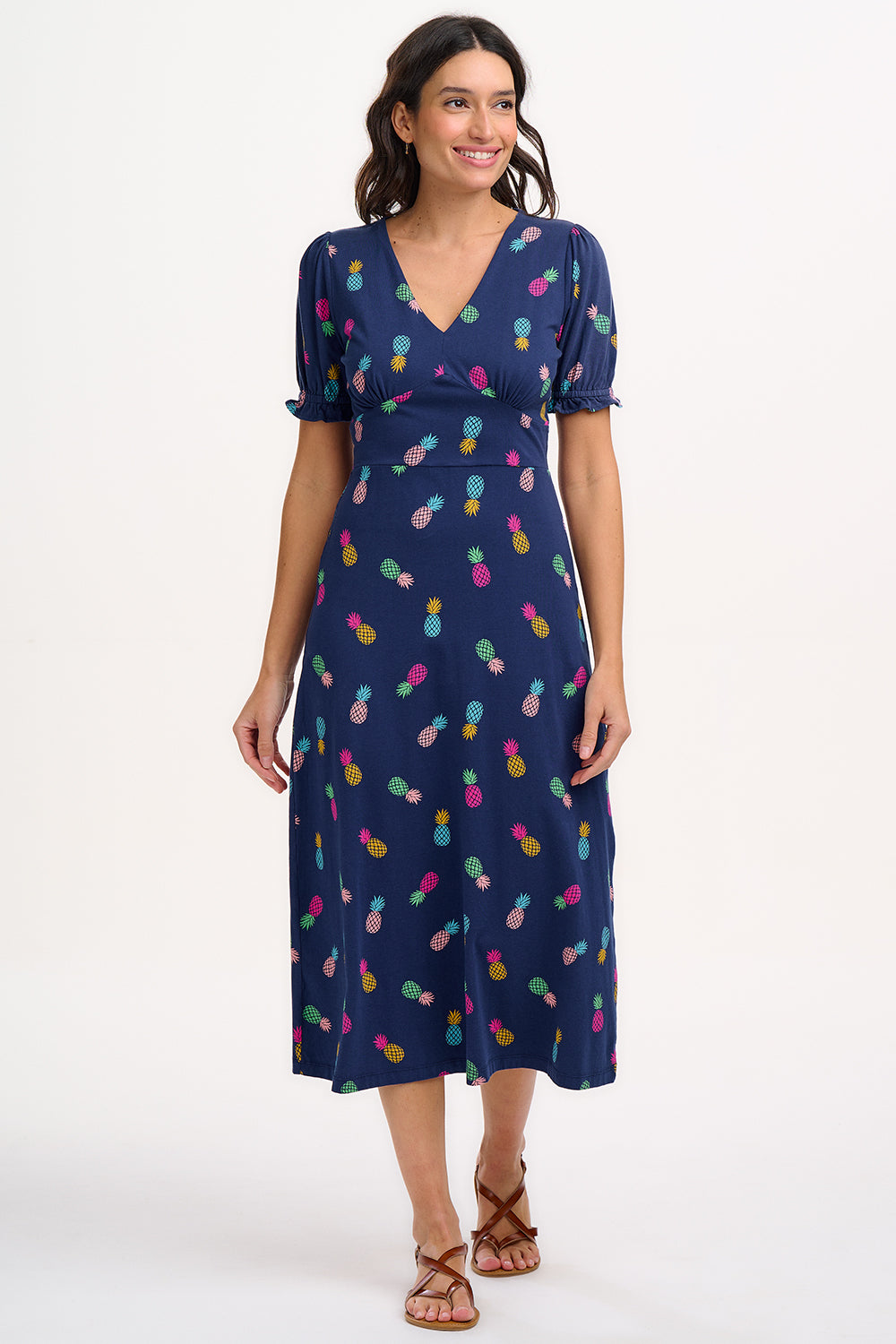 A navy midi tea dress with a multicolor pineapple print, featuring a deep V-neck and short frill sleeves, worn by a model standing against a white background.