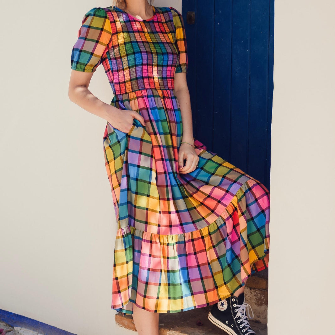 A person wearing a multicolored checkered midi dress with puff sleeves and a shirred bust, paired with sneakers, standing in front of a blue door.