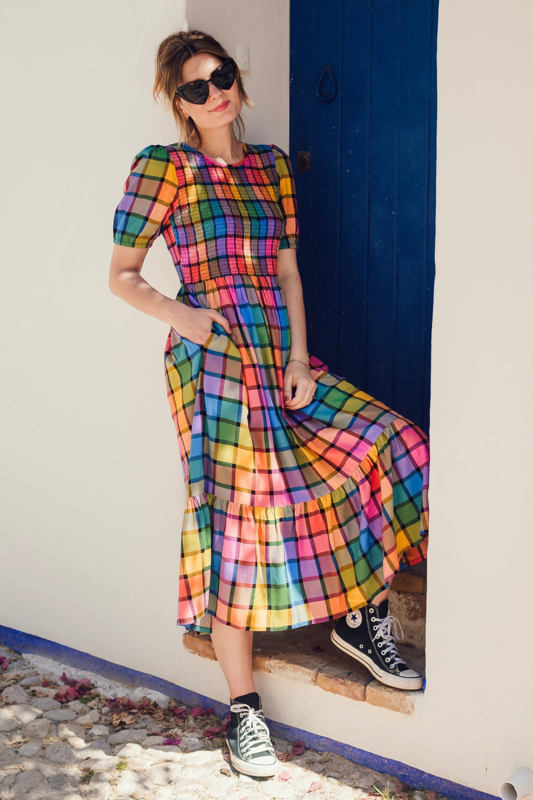 A person wearing a multicolored checkered midi dress with puff sleeves and a shirred bust, paired with sneakers, standing in front of a blue door.