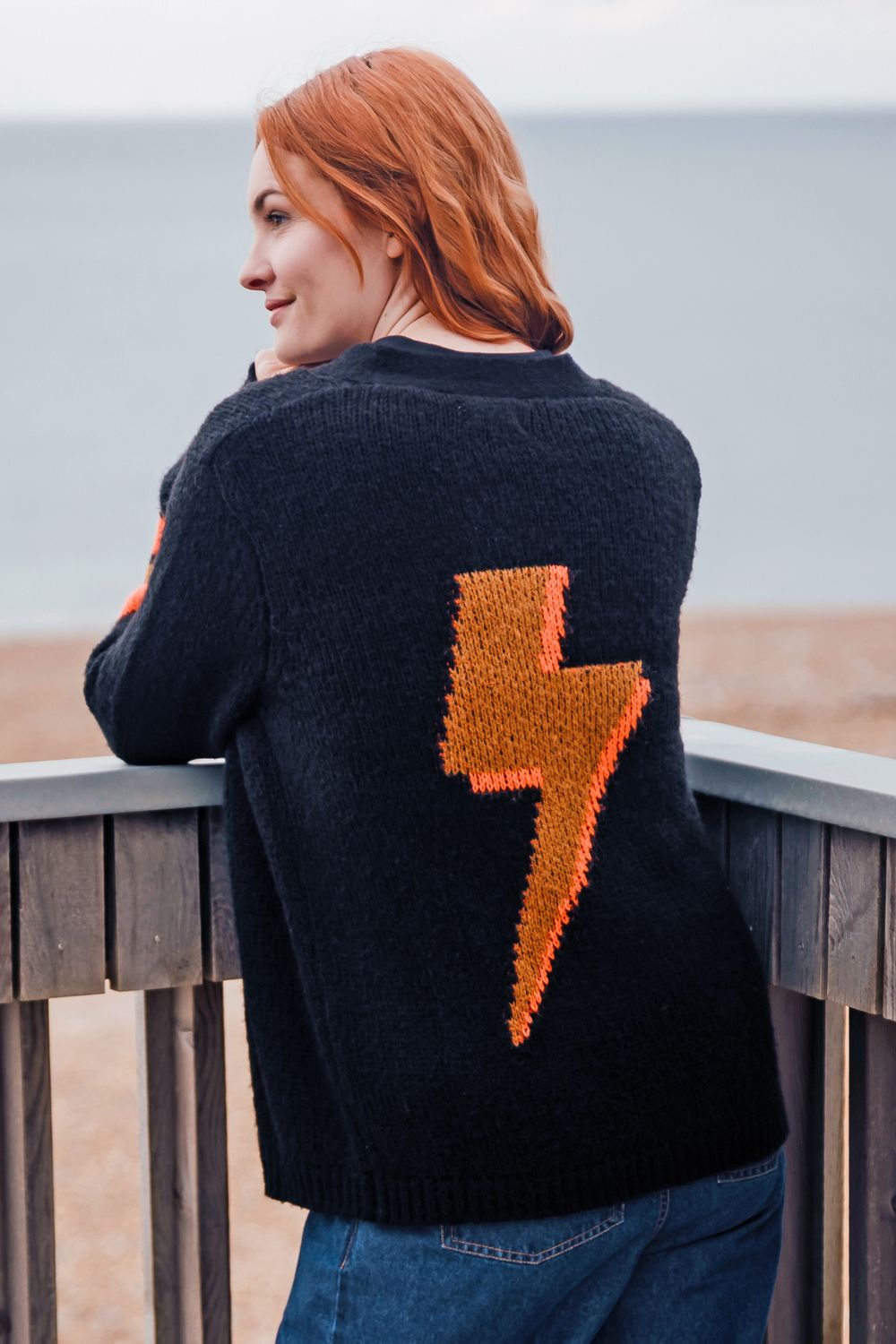 Person wearing a black sweater with an orange lightning bolt design, standing on a wooden deck by the sea.