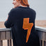Person wearing a black sweater with an orange lightning bolt design, standing on a wooden deck by the sea.