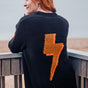 Person wearing a black sweater with an orange lightning bolt design, standing on a wooden deck by the sea.