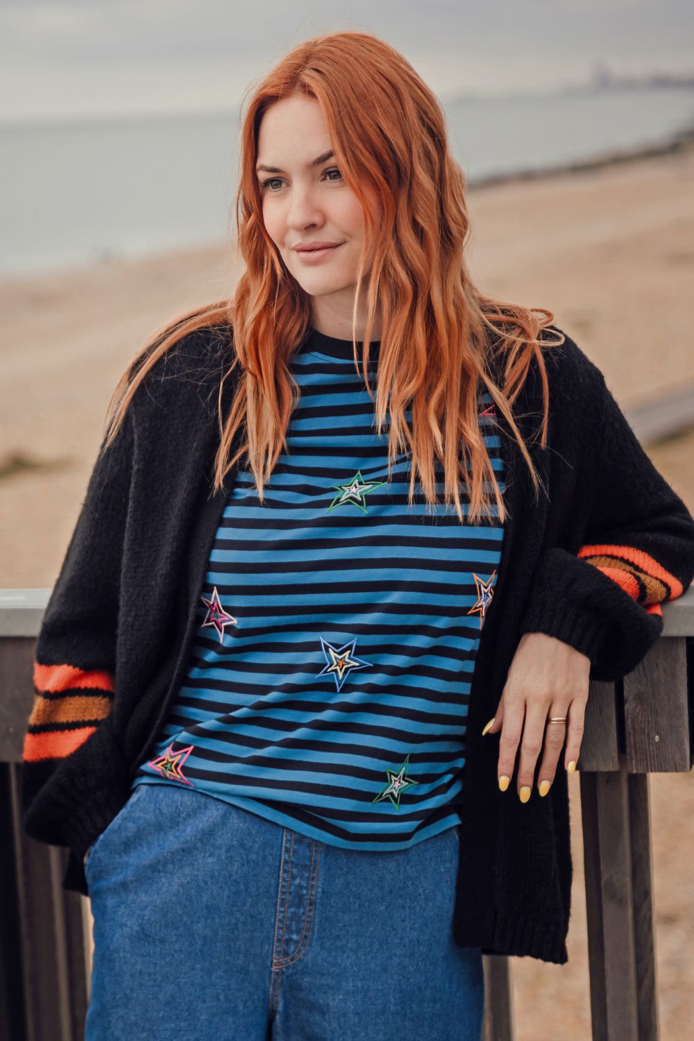 Woman with red hair wearing a black cardigan, blue striped shirt, and jeans standing outdoors.