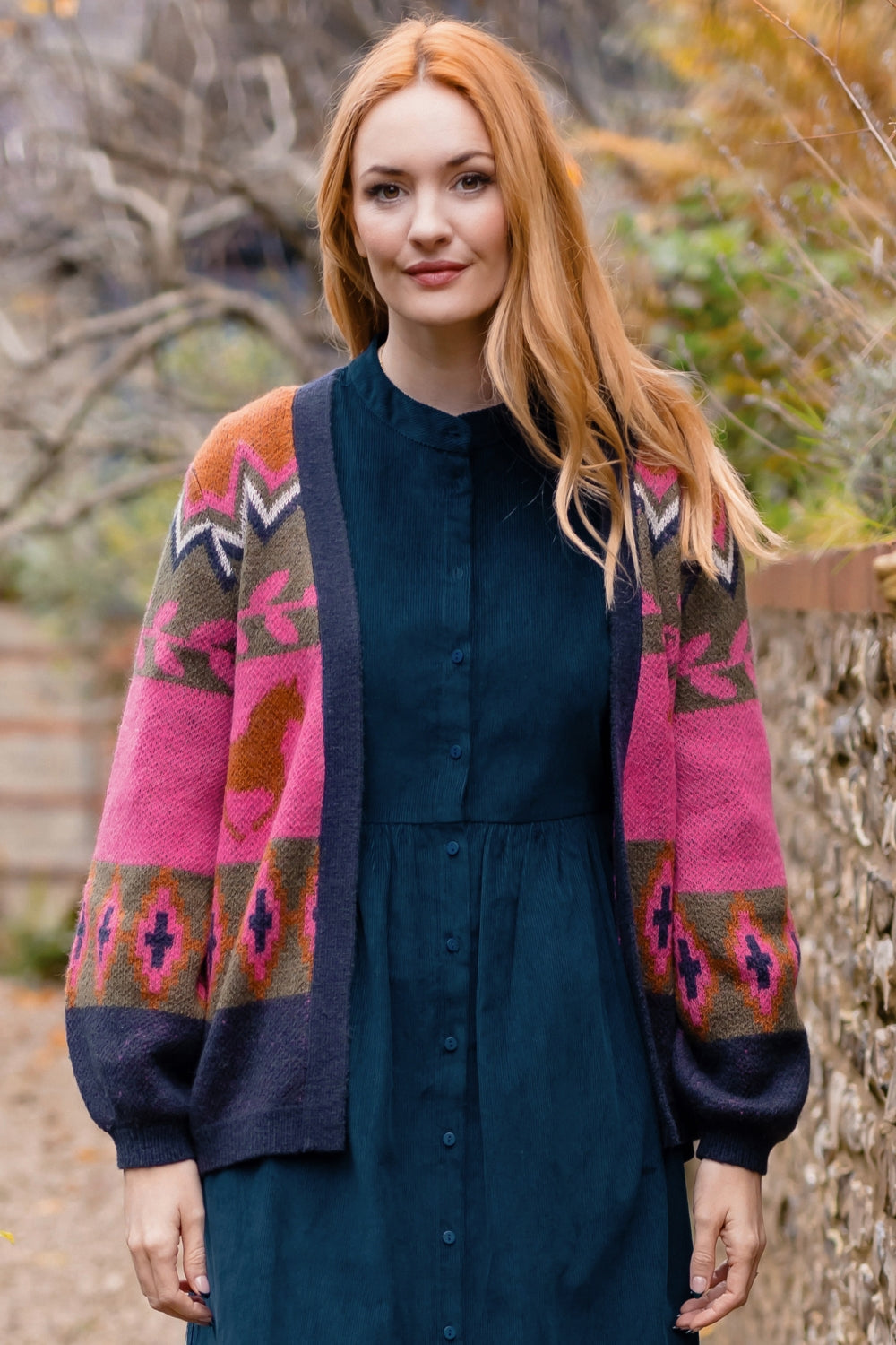 A woman with long red hair stands outdoors wearing a dark teal button-front dress layered under a vibrant pink, navy, and brown patterned knit cardigan.