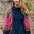 A woman with long red hair stands outdoors wearing a dark teal button-front dress layered under a vibrant pink, navy, and brown patterned knit cardigan.