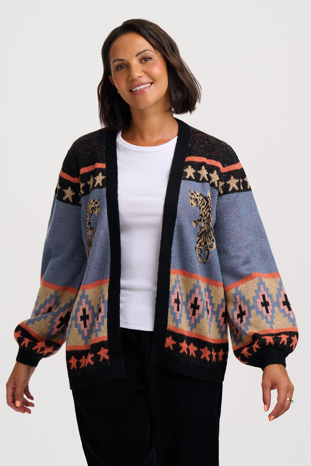 A woman wearing a black open-front cardigan with a colorful geometric and animal pattern, layered over a white t-shirt and black pants, against a light gray studio background.