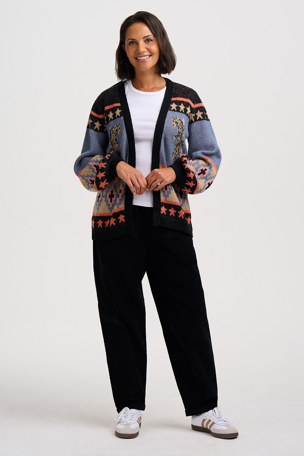 A woman modeling a black and blue patterned cardigan with star and geometric motifs, worn over a white t-shirt and black pants, on a light gray studio background.